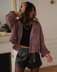 Veste en jean prune Suki à pressions argentées, portée ouverte avec un short noir, une main dans les cheveux